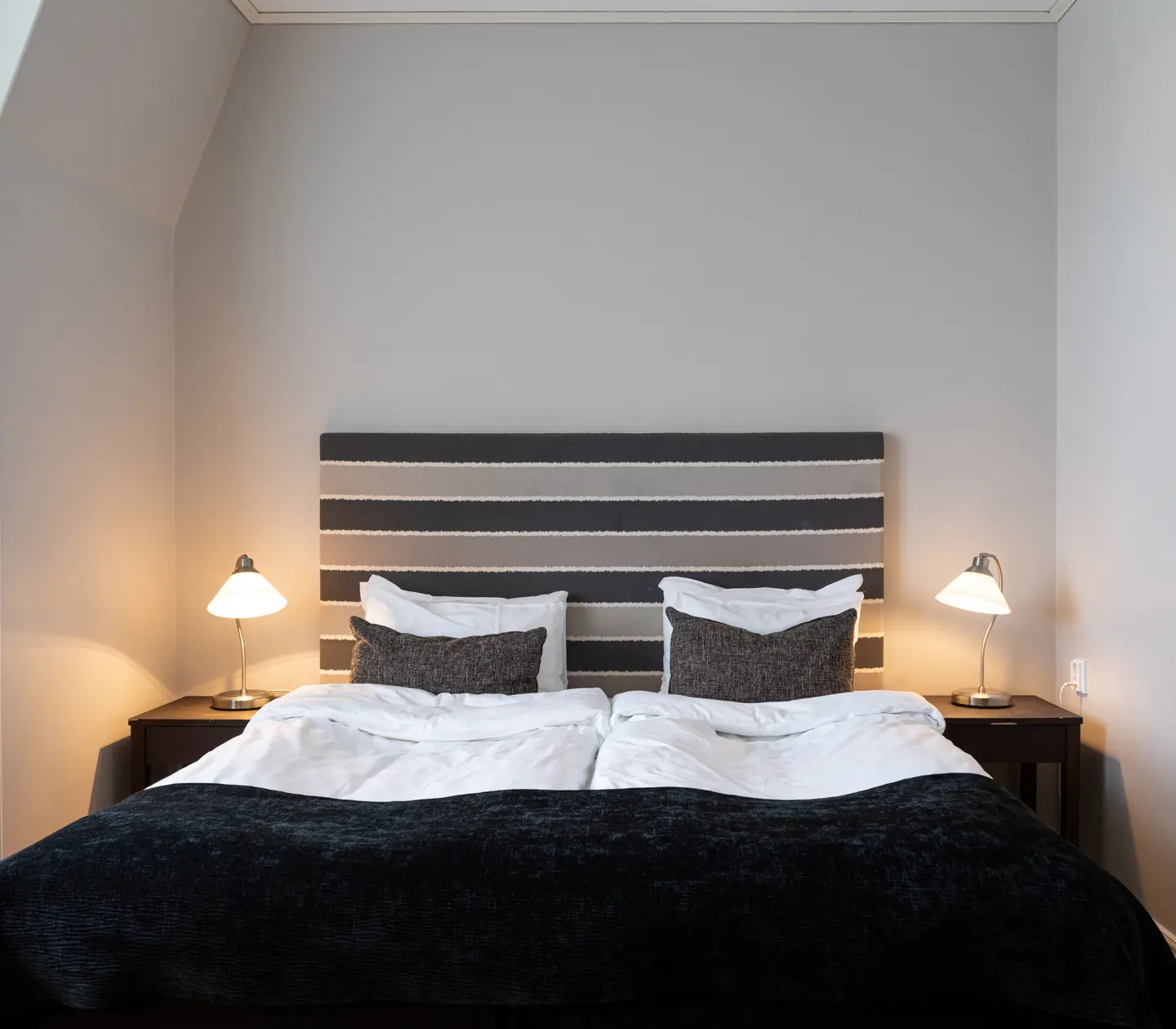 Modern hotel room with a double bed featuring striped headboard, white bedding, and dark throw blanket.