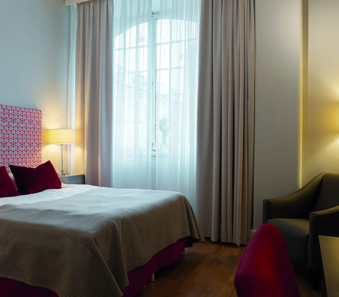 Modern hotel room with a plush bed featuring a patterned headboard, accented by red pillows and a beige throw, beside a window with sheer curtains allowing natural light.