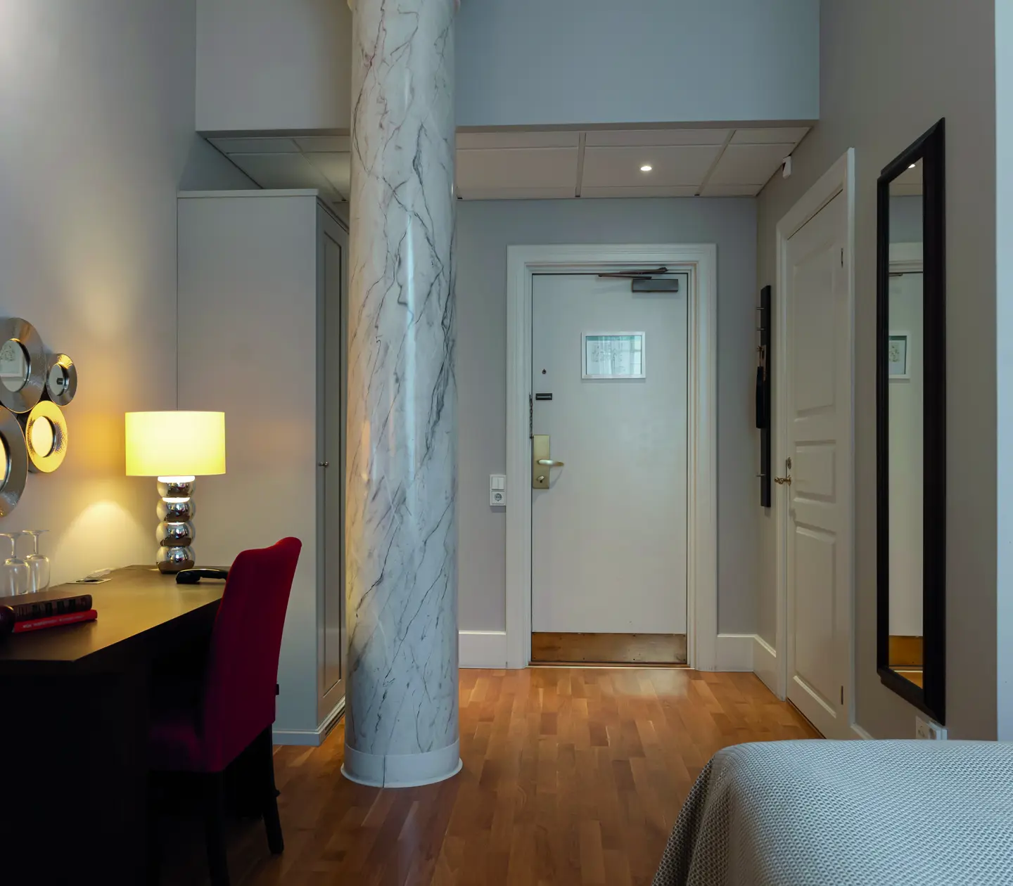 Contemporary hotel room with a chic marble column, elegant wall mirrors, a sleek desk with a stylish lamp, and a bright red chair.