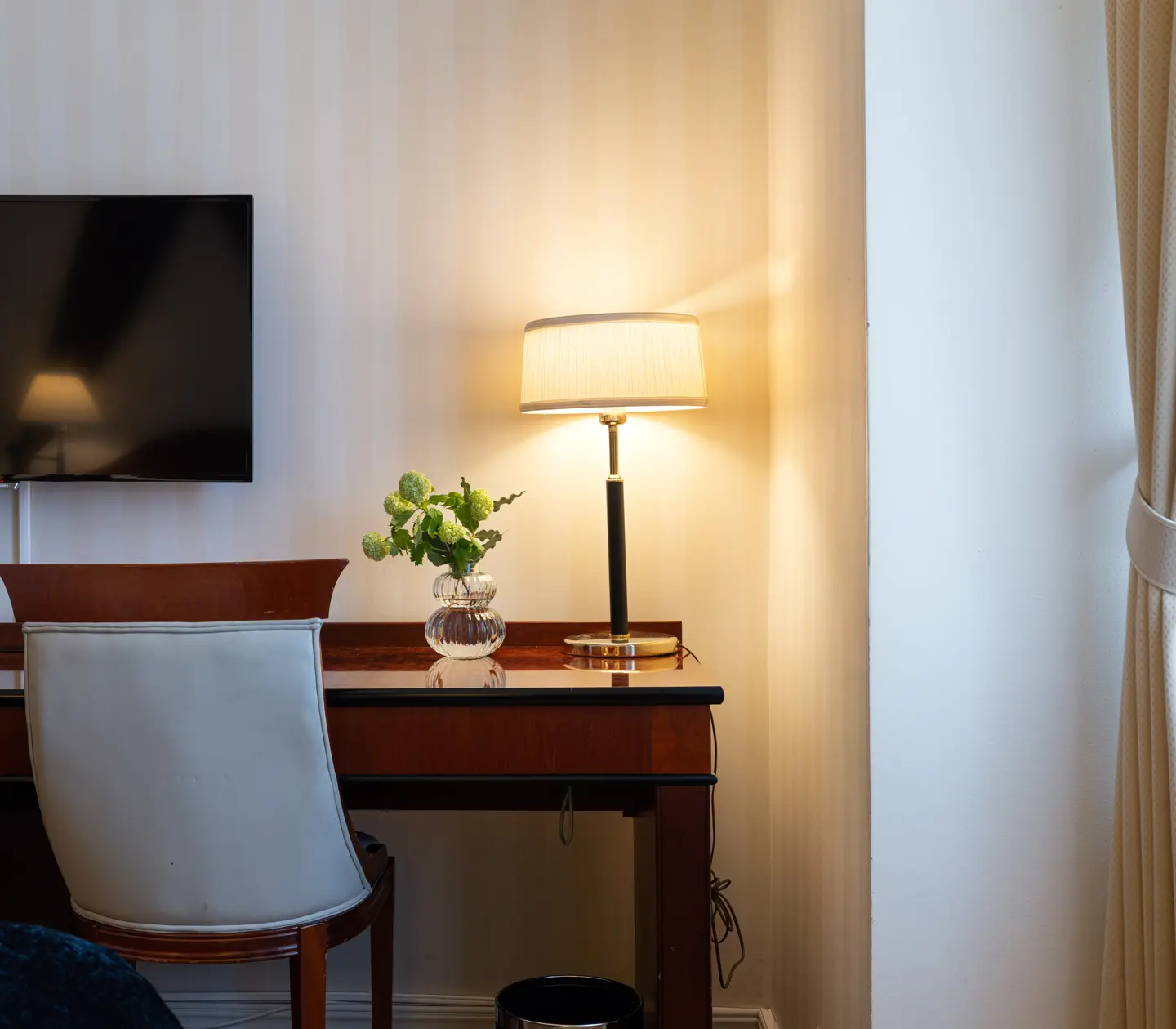 Hotel room interior with a wooden desk, beige chair, and table lamp. A TV is mounted on the wall above the desk, and a vase with green flowers is on the surface.