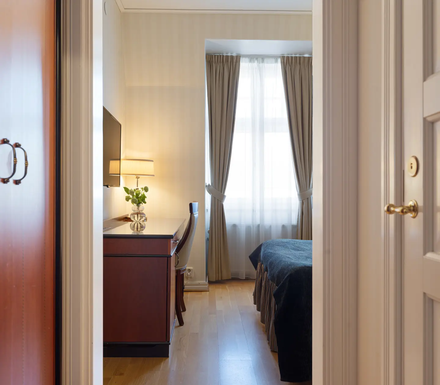 Cozy hotel room with a neatly made bed, wooden furniture, and a large window with curtains letting in natural light.