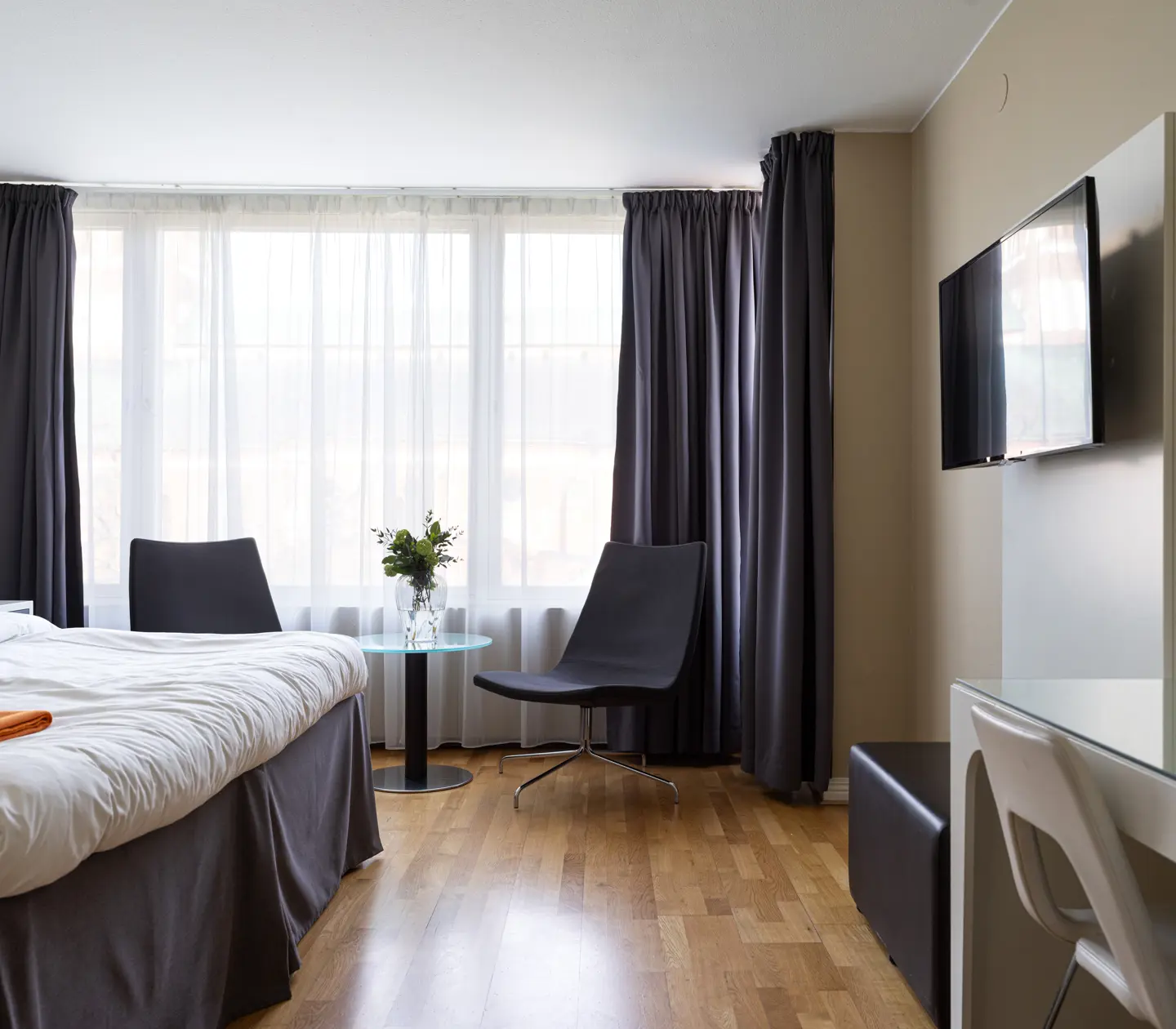 Modern hotel room with a comfortable bed, two black chairs, a glass table with flowers, a wall-mounted TV, and large windows with gray curtains.