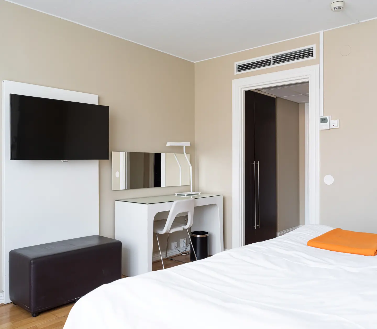 Modern hotel room interior with a wall-mounted flat-screen TV, a sleek desk with a mirror and chair, and a bed with a white duvet and orange accent.