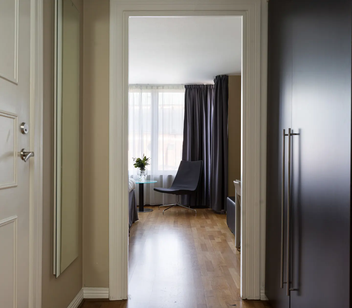 Modern hallway leading to a bright room with wooden flooring, a black chair, large window, and dark curtains.