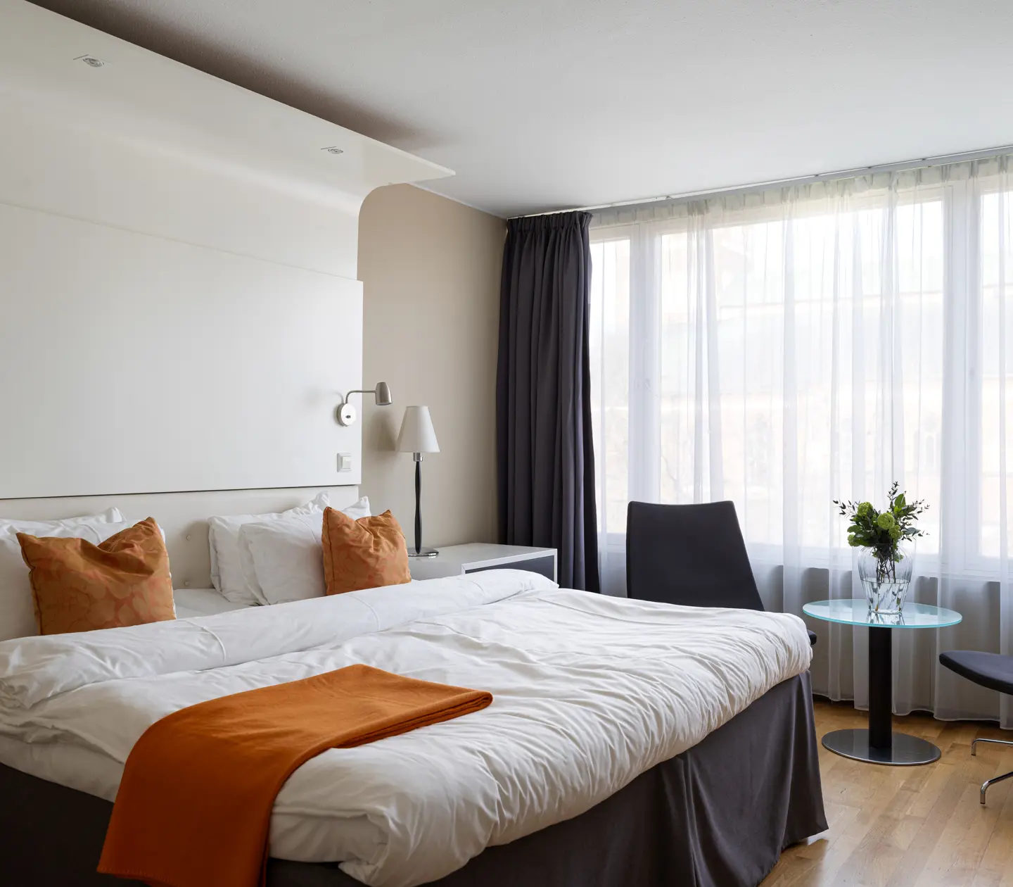 Modern hotel room with a large bed featuring white linens and orange accents, a seating area with two chairs, a small glass table with a vase of flowers, and ample natural light from large windows with sheer curtains.