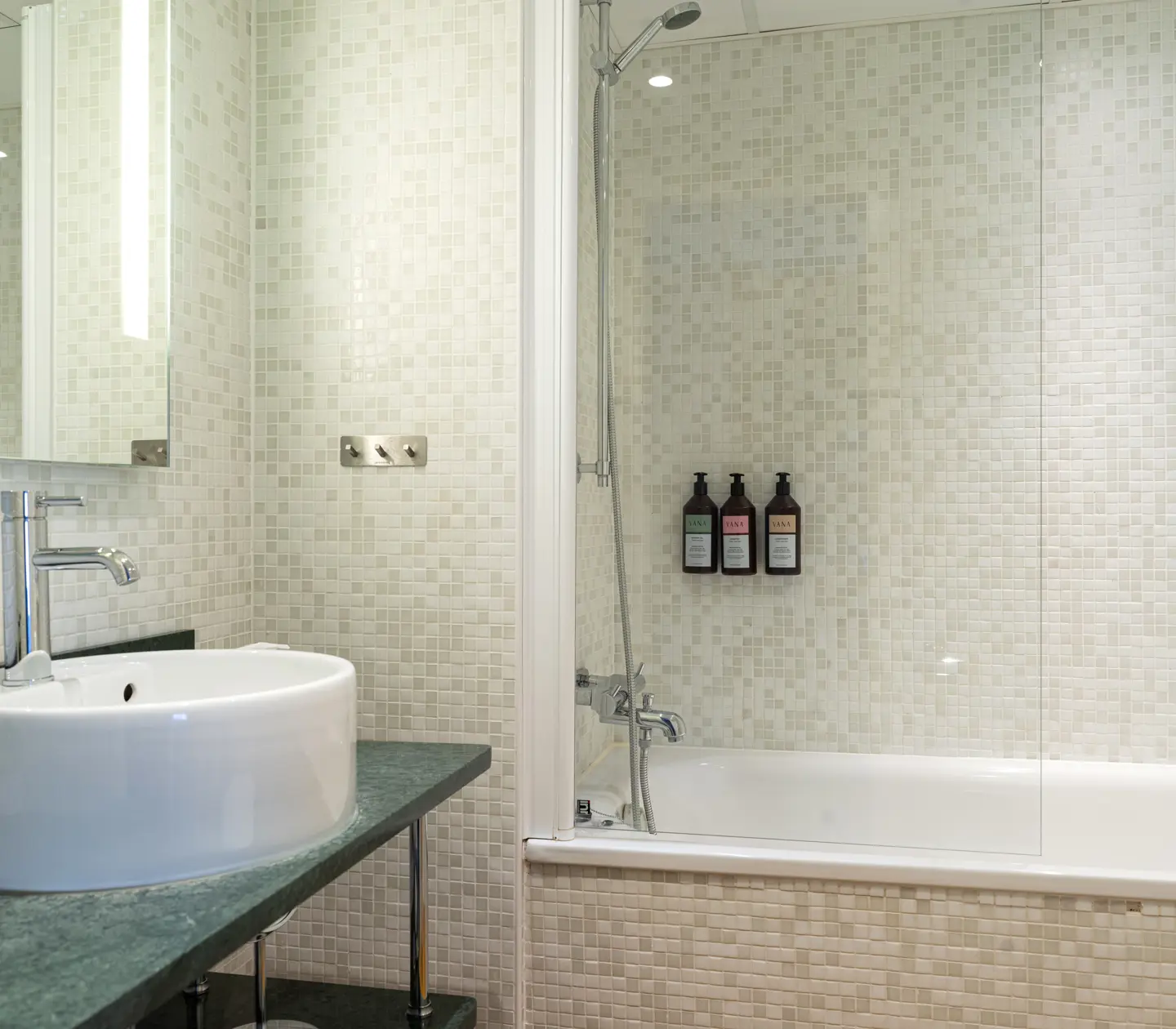 Modern bathroom with white mosaic tiles, featuring a sleek vessel sink and faucet on a dark countertop, adjacent to a bathtub with a glass shower screen and mounted toiletries.