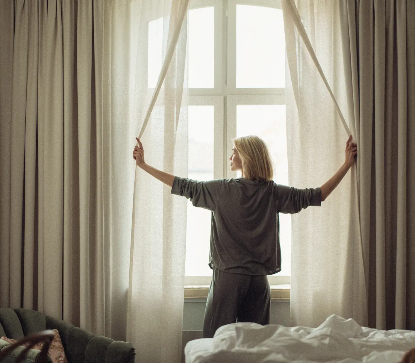 Kvinna i grått loungewear öppnar gardiner vid stort fönster, släpper in dagsljus i sovrum med säng och fåtölj.