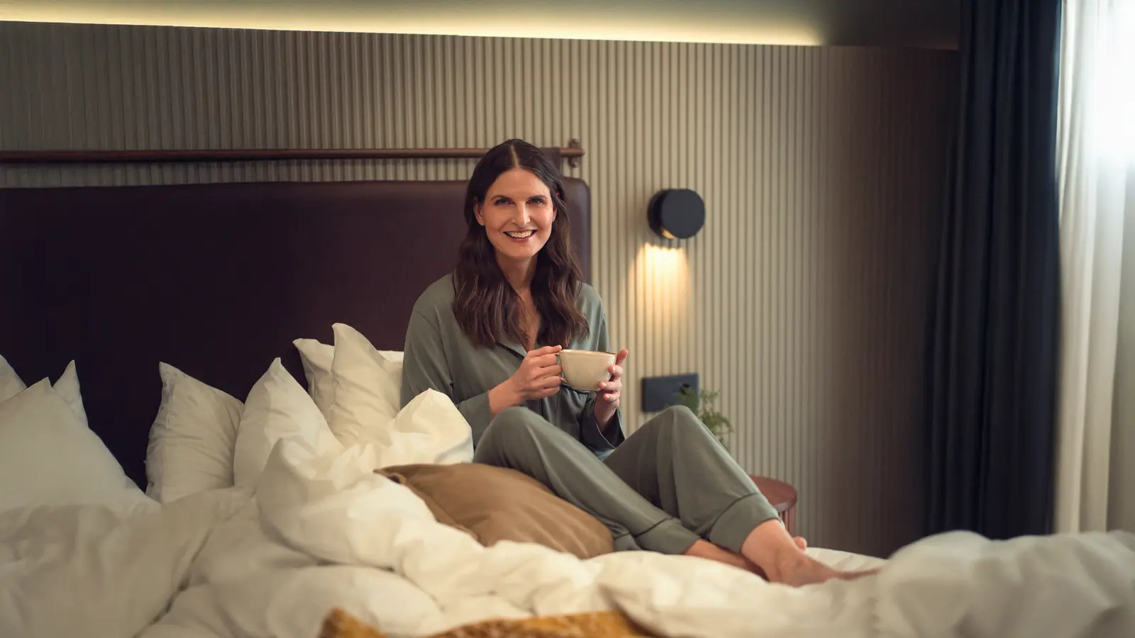 Kvinna i pyjamas sitter leende på en säng och håller en kopp, omgiven av kuddar i ett minimalistiskt sovrum.