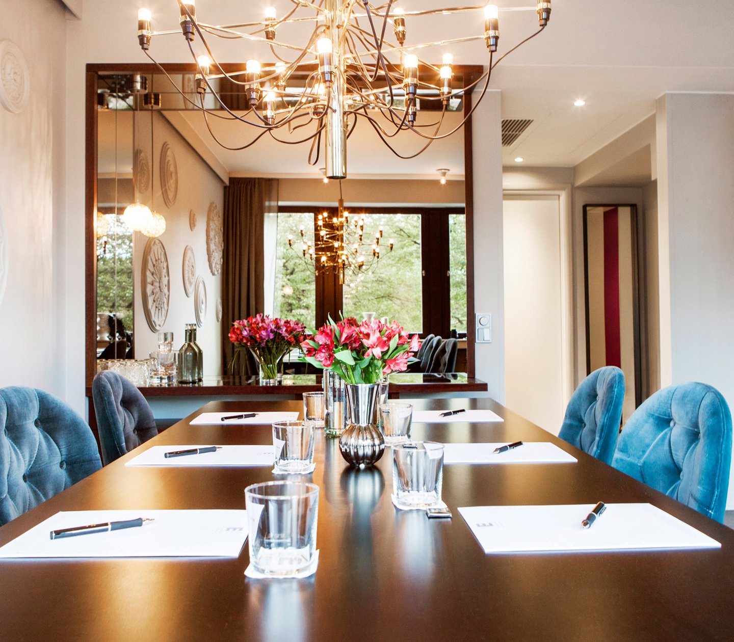 A beautiful conference room with natural lightning, greenery outside the window, blue velvet chairs and a big bouquet on the table