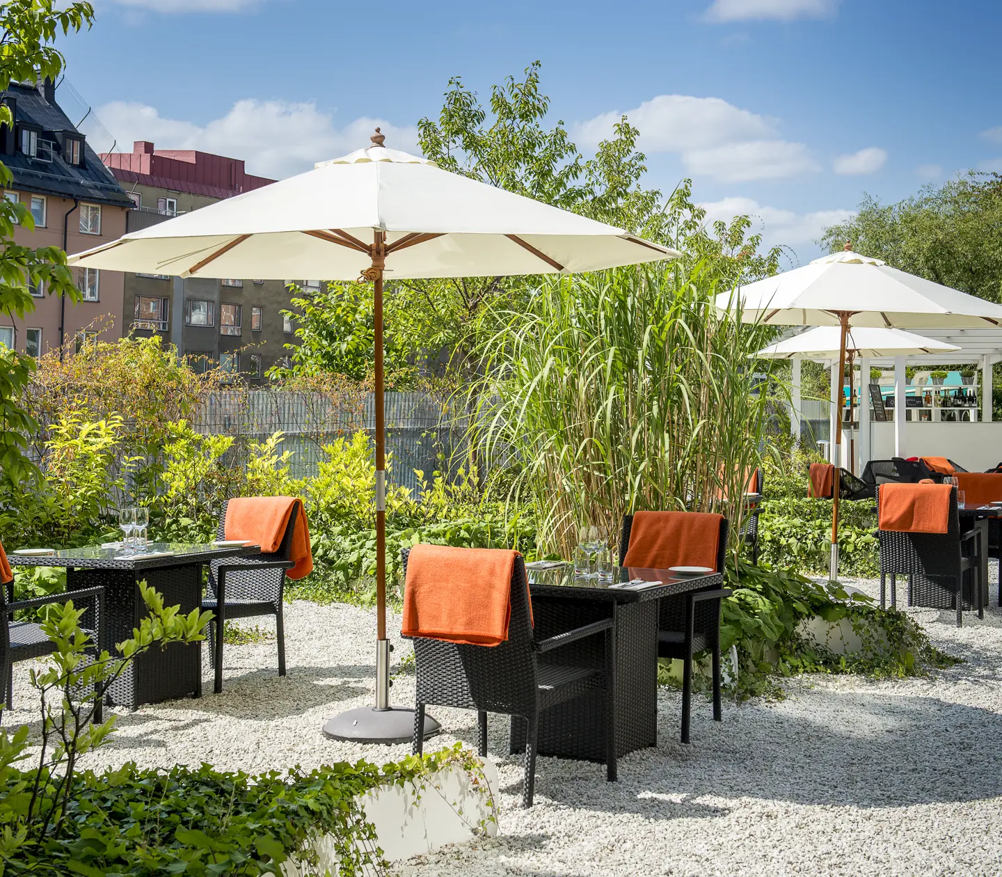 Uteservering i trädgårdsmiljö med parasoller och svarta caféstolar med orange tyg, omgiven av grönska och flerfamiljshus i bakgrunden under en klarblå himmel