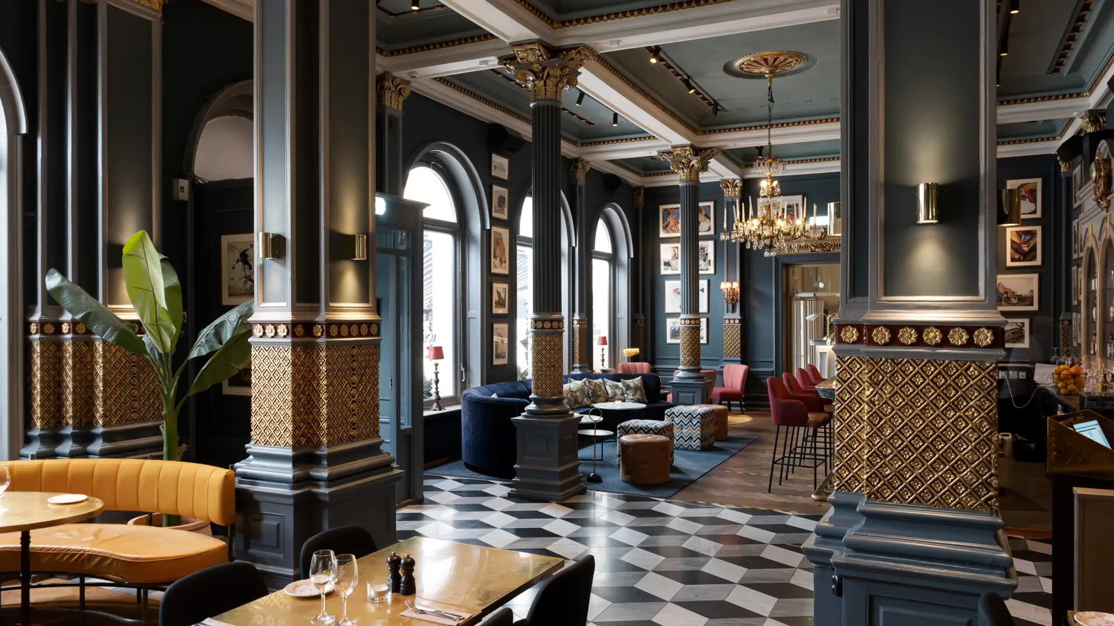 Elegant hotel lobby with classic decor, featuring ornate column details, plush seating, chandeliers, and framed artwork. The space is well-lit with large windows and a patterned tile floor, creating a luxurious and inviting atmosphere.