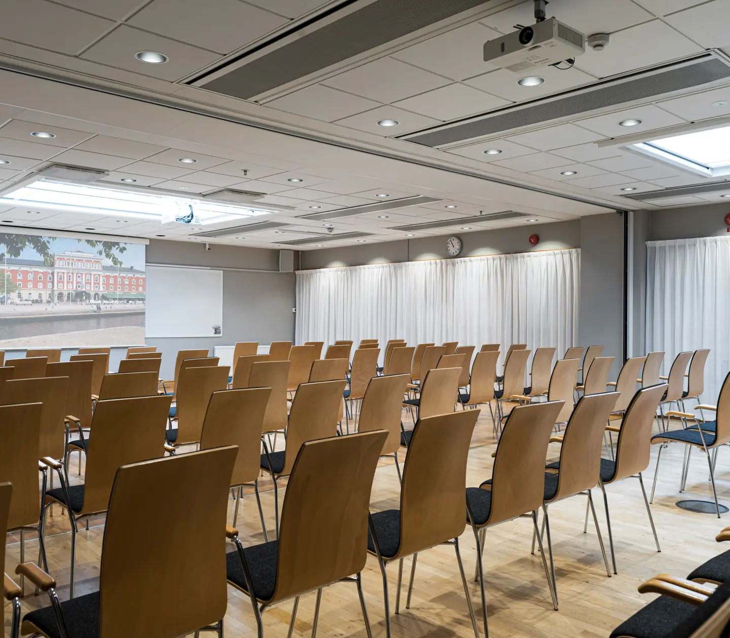 Konferensrum med projektor och rader av trästolar, redo för presentation eller möte. Ljusa gardiner och vitt tak med LED-belysning.