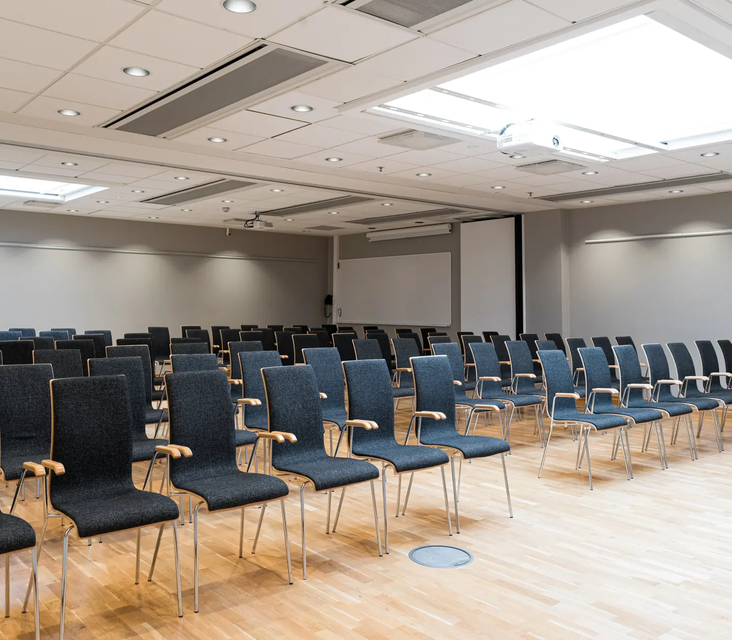 Konferensrum med rader av tomma stolar, parkettgolv och projektor i taket.