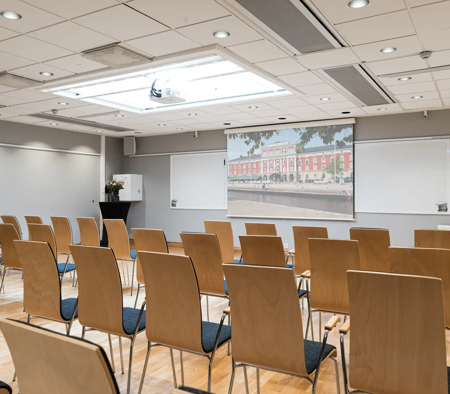 Modernt konferensrum med trästolar och projektorduk, belyst av naturligt ljus från stora takfönster.