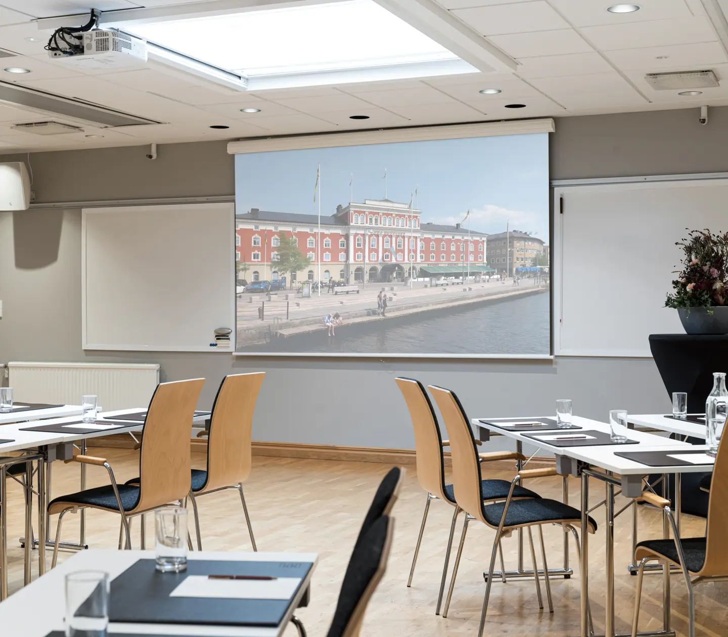 Konferensrum med projektor som visar bild av ett historiskt byggnad vid vattnet i Sverige. Bord med stolar, anteckningsmaterial och vattenflaskor.