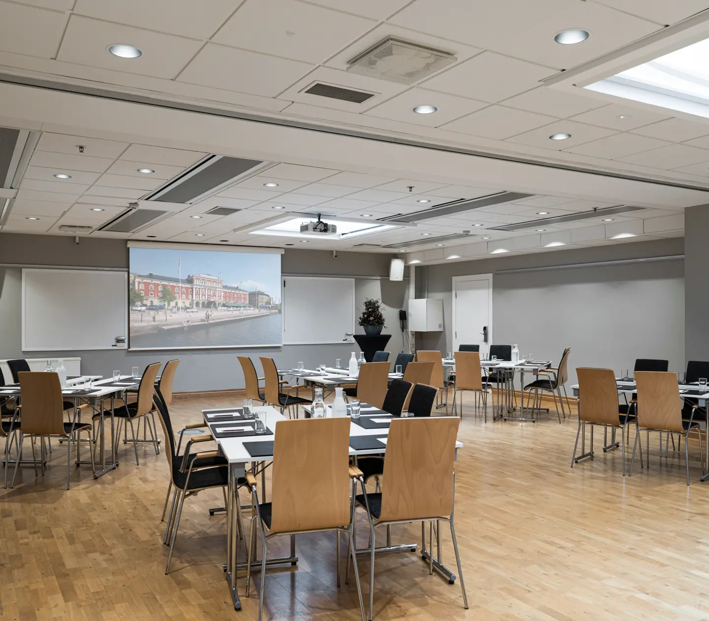Konferensrum med trägolv och moderna möbler, redo för presentation med projektorduk i framkant.