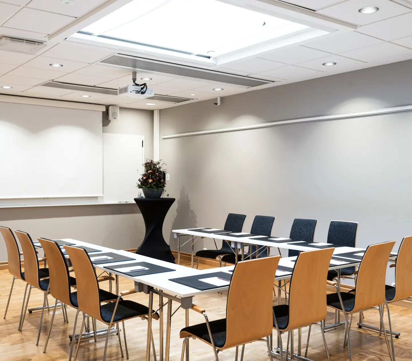 Konferensrum med U-format bord, stolar, projektor och projektorduk. Passar för möten och presentationer.