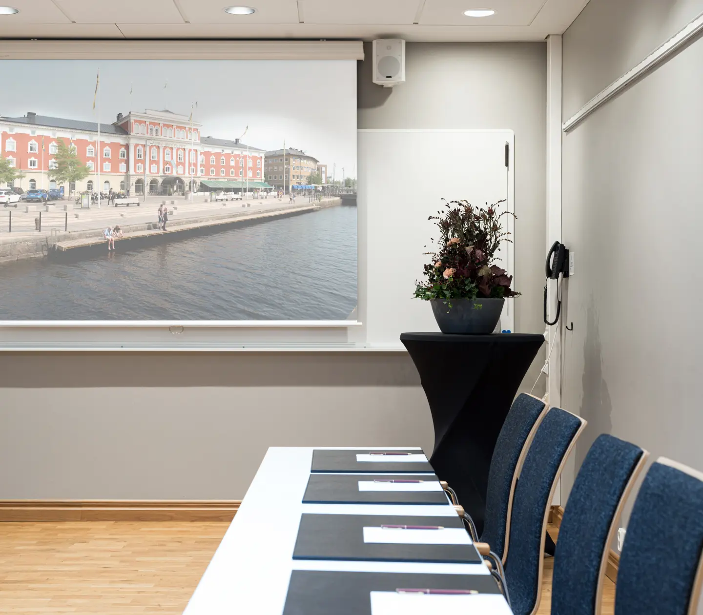 Konferensrum med projektor som visar en bild av en historisk byggnad vid vattnet, bord med stolar och blommor på ett sidobord.
