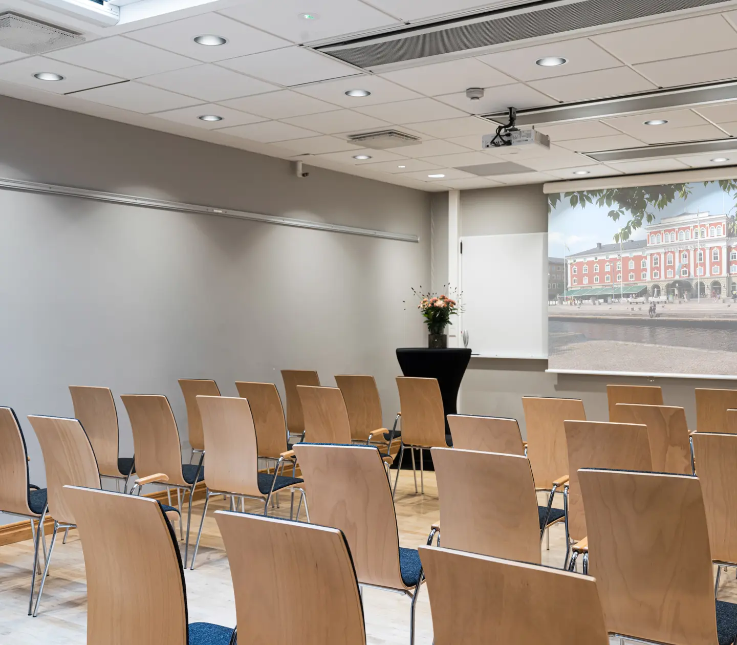 Konferenslokal med trästolar och en projicerad bild av en historisk byggnad på duken. Ljusa väggar och ett arrangemang av blommor vid en talarstol.