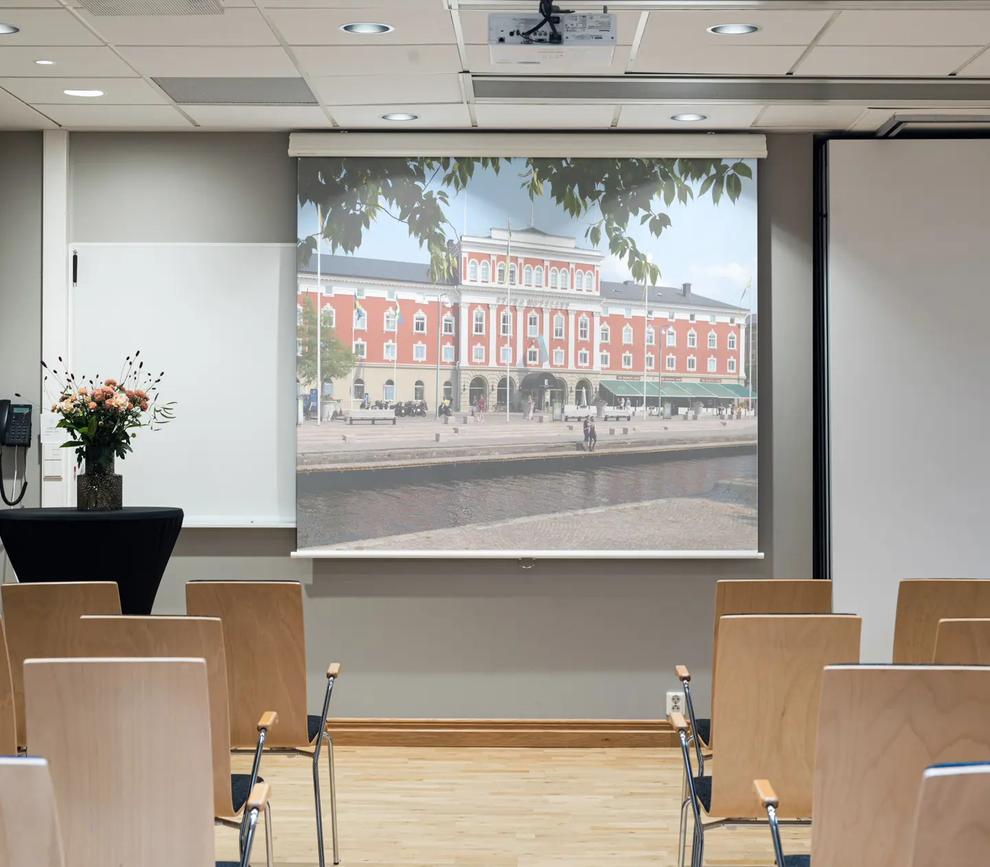 Konferensrum med projicerad bild av en historisk byggnad i rött och vitt, troligen belägen vid en kanal. Bänkrader av trästolar, blombukett på bord.