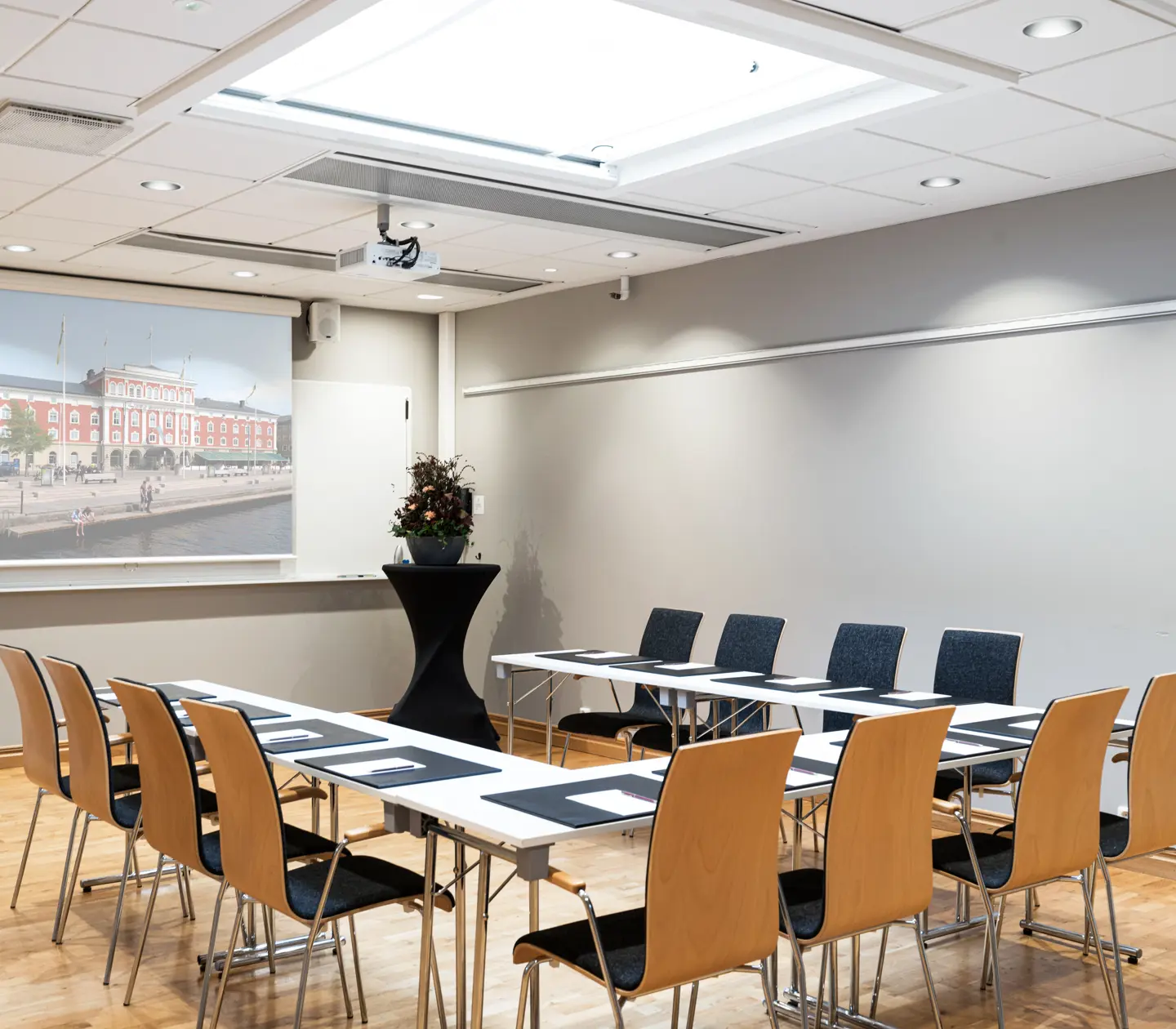 Konferensrum med projektor, duk och sittplatser arrangerade i U-form, trästolar och bord, modern inredning, lämpligt för möten och presentationer.