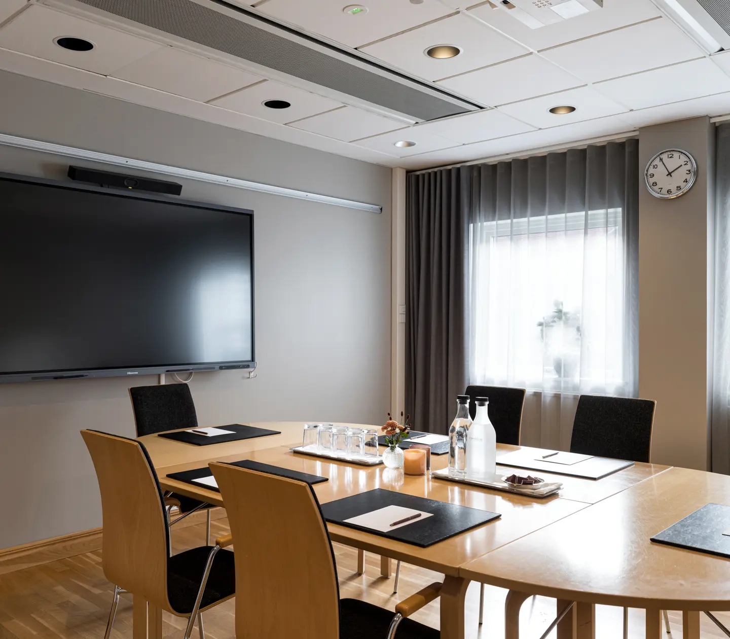 Modernt mötesrum med stor skärm, träbord och svarta stolar, förberett för konferens med vattenflaskor och anteckningsblock.
