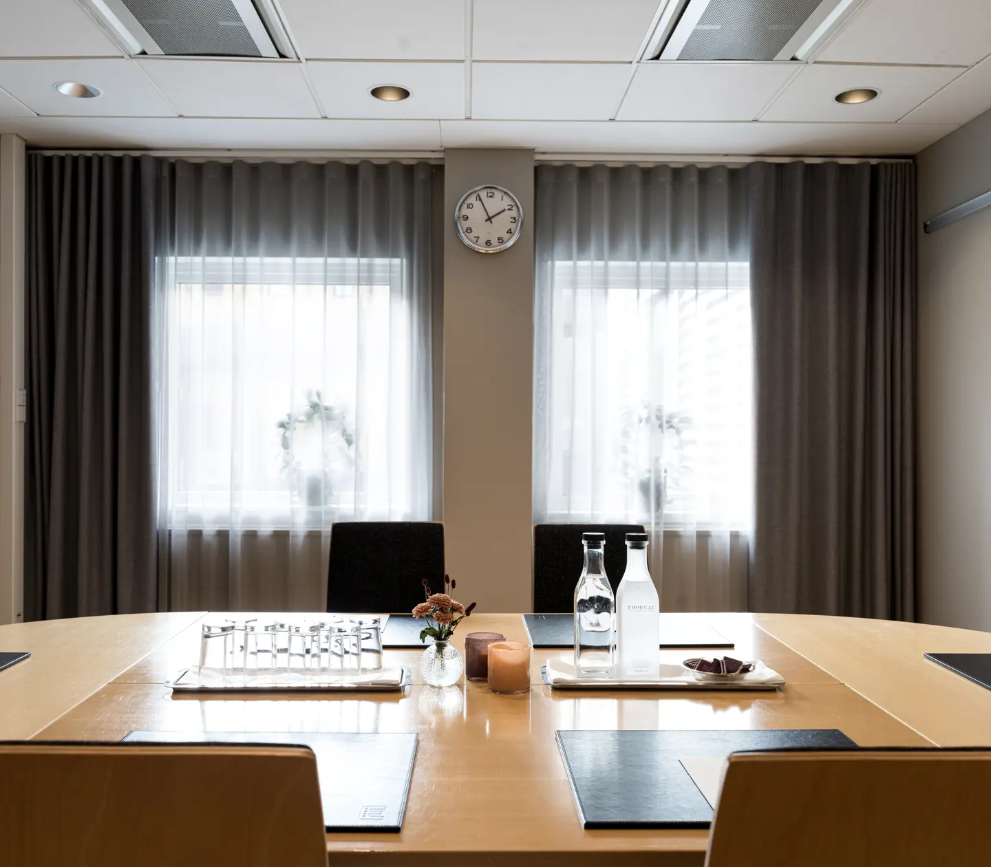 Modernt konferensrum med ovalt bord, glasflaskor och dekorationer; klocka på väggen mellan två fönster med gardiner.