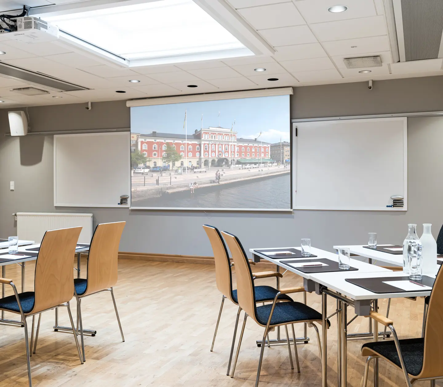 Konferensrum med projektor och sittplatser, visande en bild av ett historiskt byggnad vid vattnet. Ljusa väggar och trägolv skapar en modern och professionell miljö för möten och presentationer.