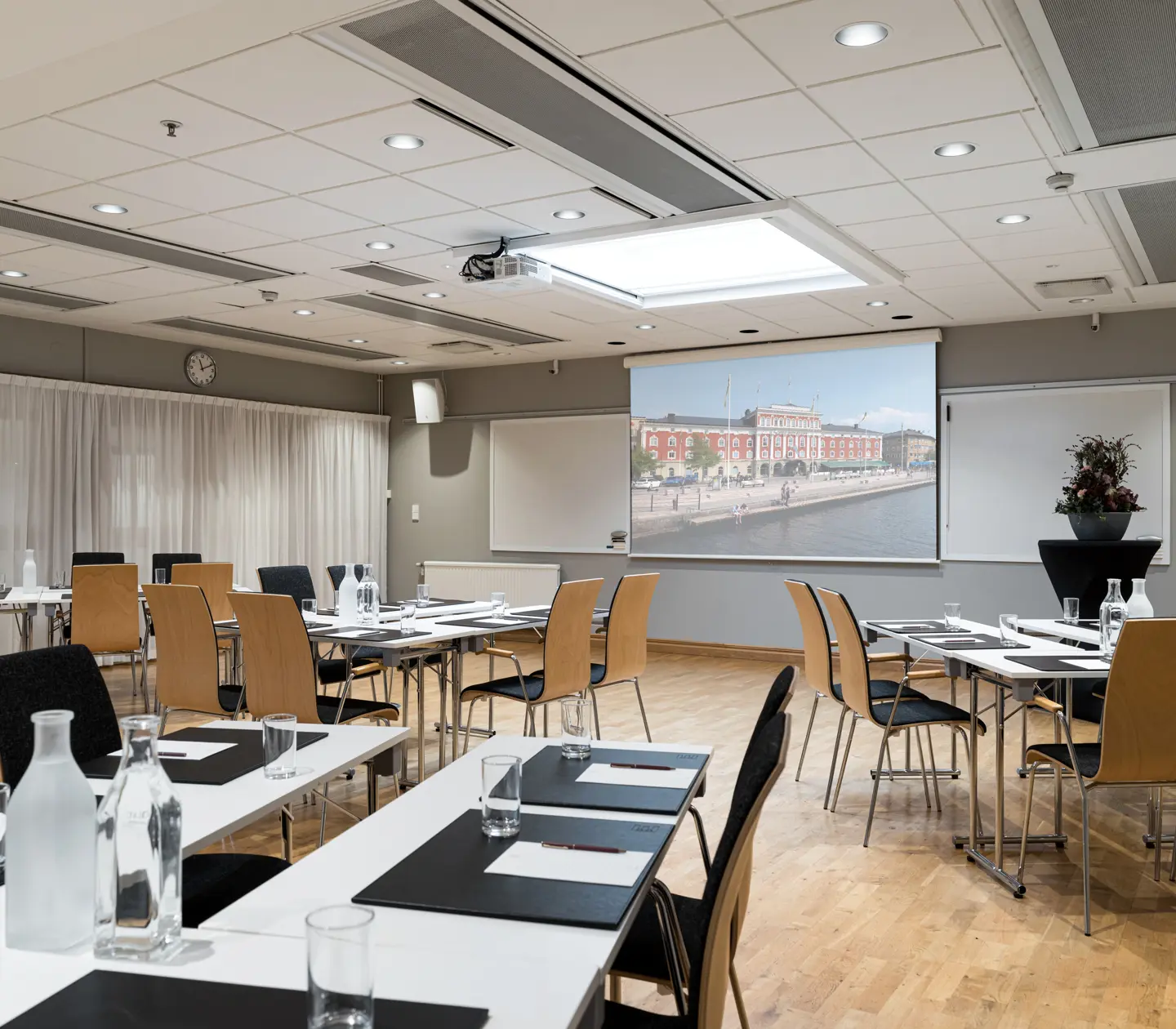Konferensrum med uppställda bord och stolar, presentationsskärm med stadsvy i bakgrunden, väl upplyst och modernt inrett för affärsmöten.