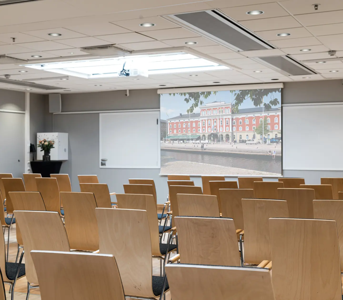 Konferensrum med trästolar, projektorduk och tavlor. Ljusa väggar och blommor på ett bord.