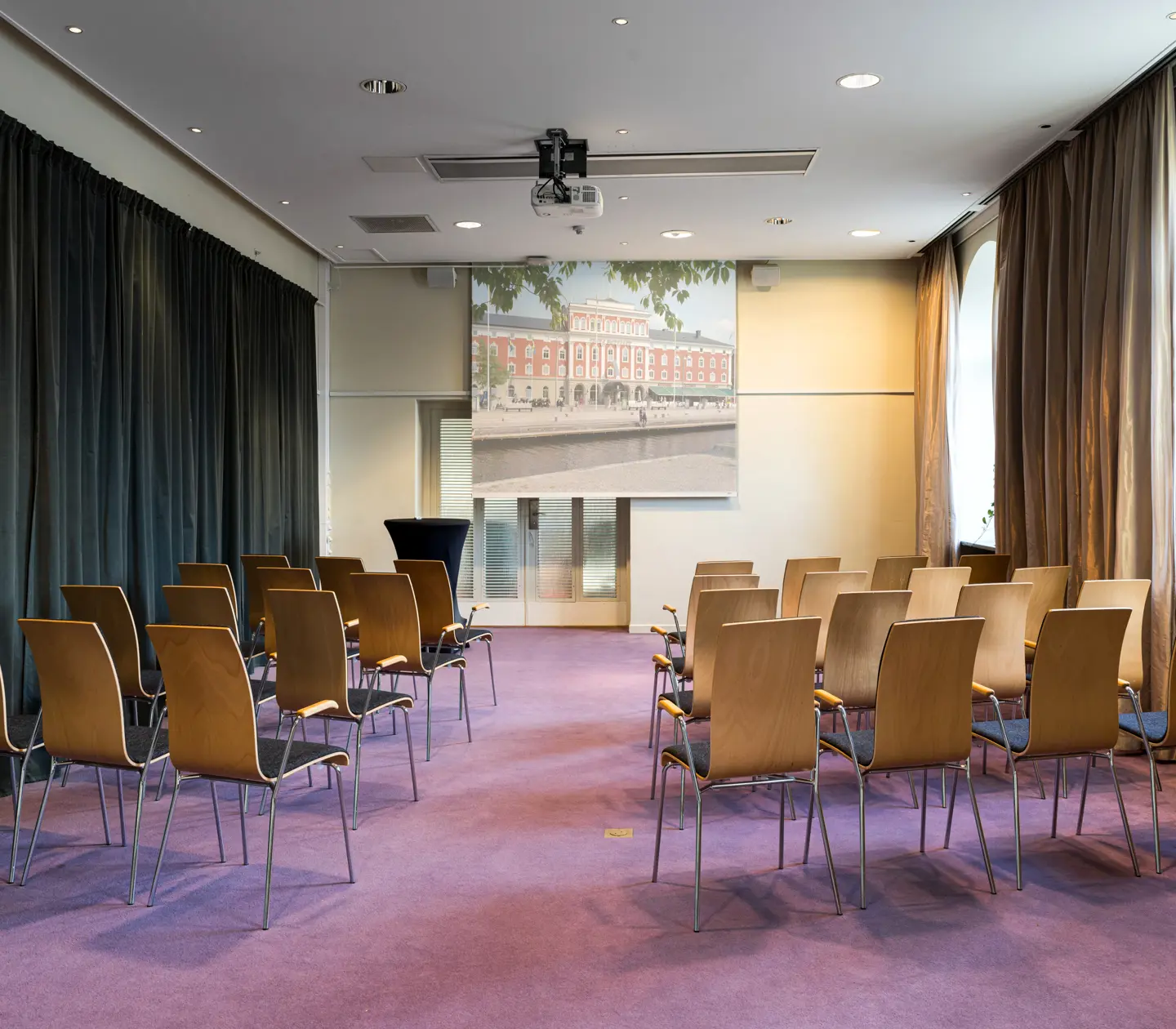 Konferensrum med stolsrader, projektorduk och gardiner. Perfekt för möten och presentationer.