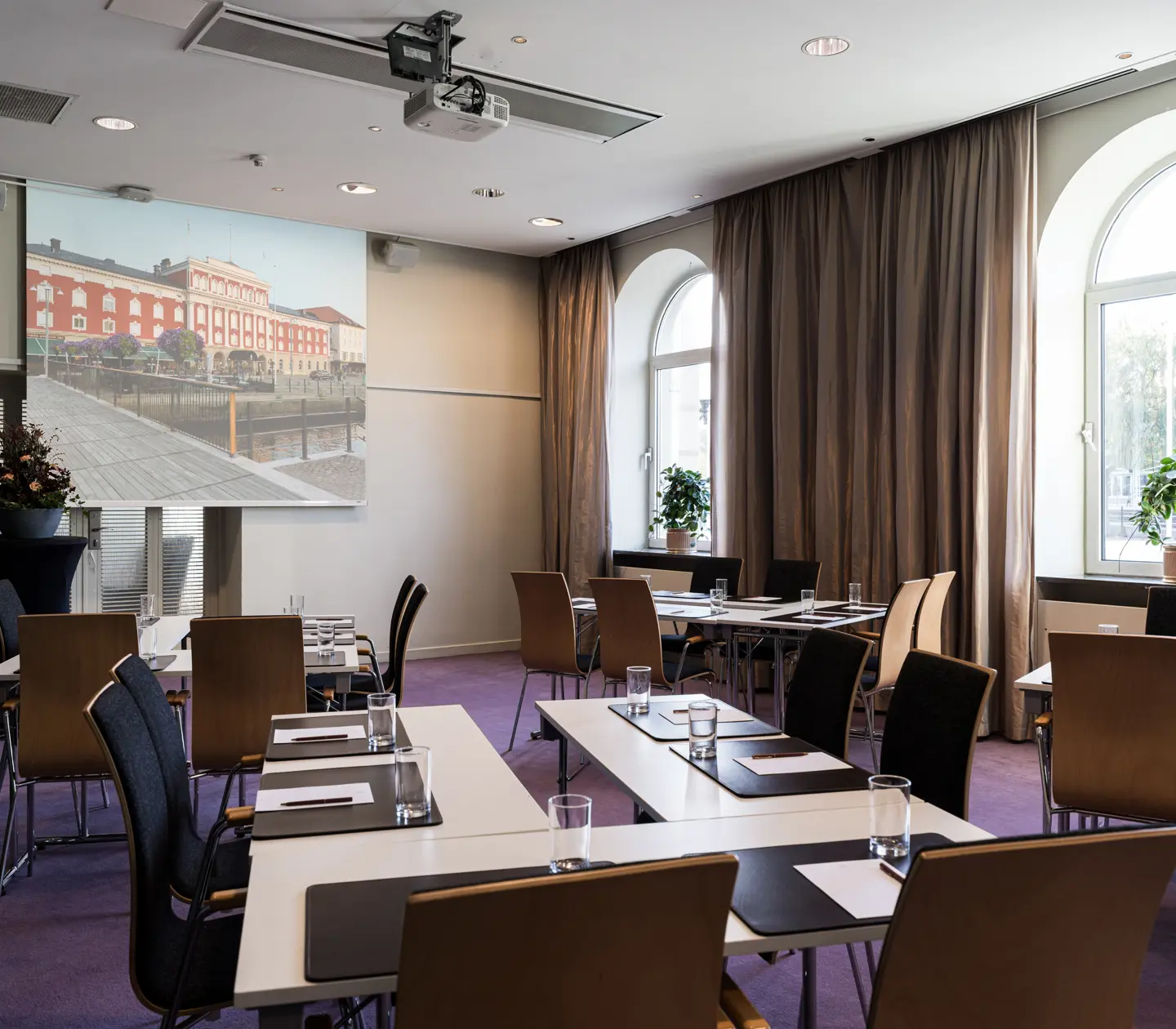 Modern konferensrum med projektorskärm och fönster, utrustat med skrivbord och stolar, idealiskt för möten och presentationer.