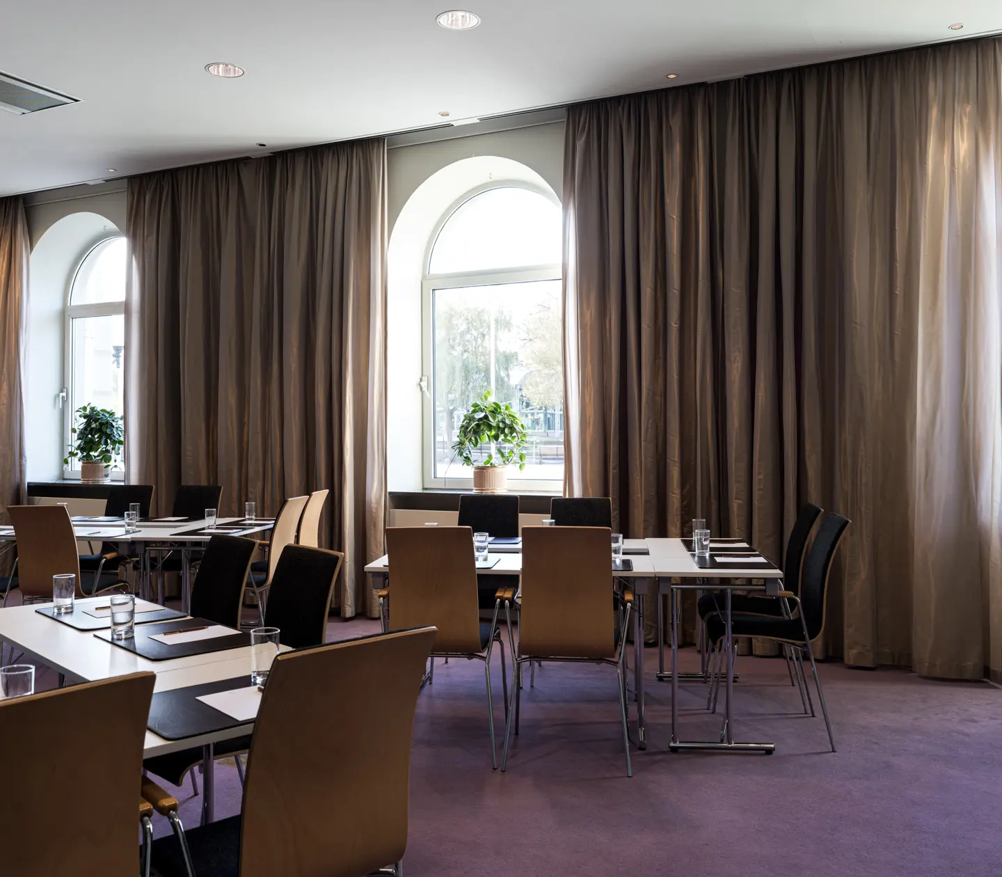 Konferensrum med uppställda bord och stolar, stora fönster med gardiner och krukväxter. Naturligt ljus strömmar in i rummet och skapar en inbjudande atmosfär.