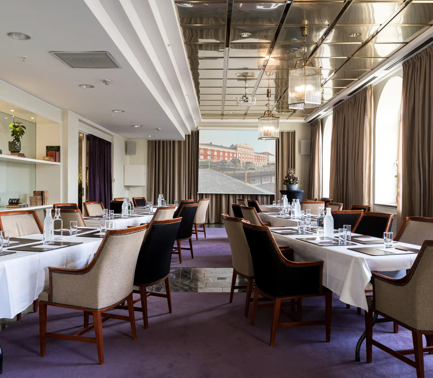Konferensrum med uppsättning av bord och stolar, vit dukning och projektor i en elegant miljö med stora fönster och lila matta.