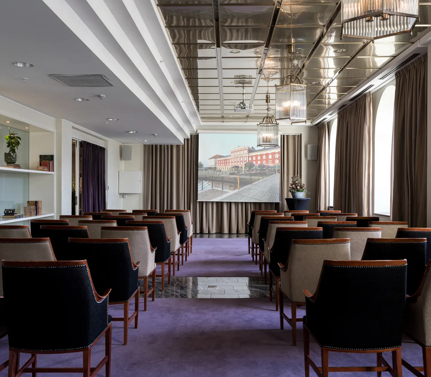 Konferensrum med uppställda stolar och projektorduk, elegant inredning med modernt tak och stora fönster med cremefärgade gardiner.