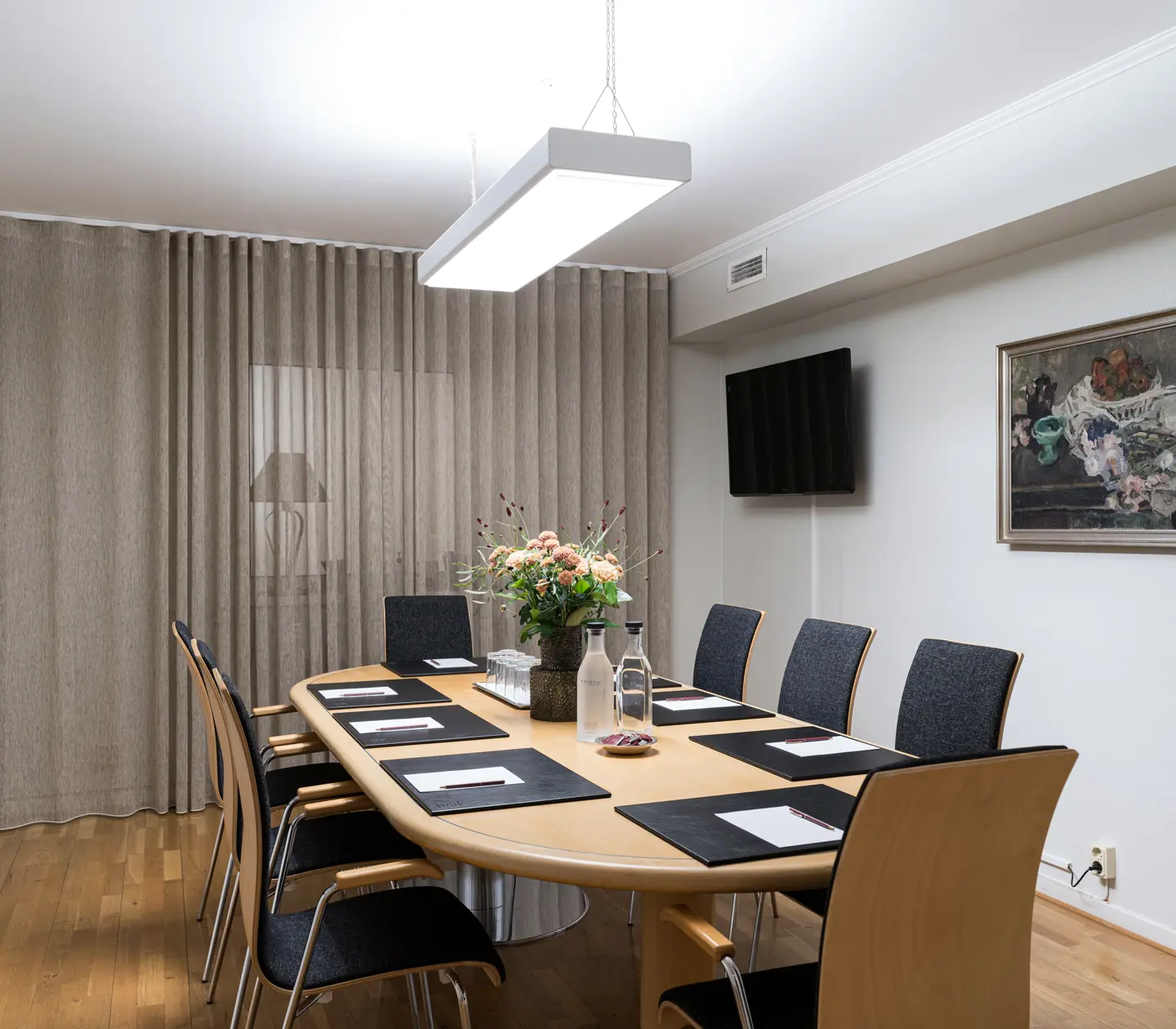 Modernt konferensrum med ett ovalt bord, åtta stolar, blommor på bordet, vägg-TV och målning, ljus interiör.