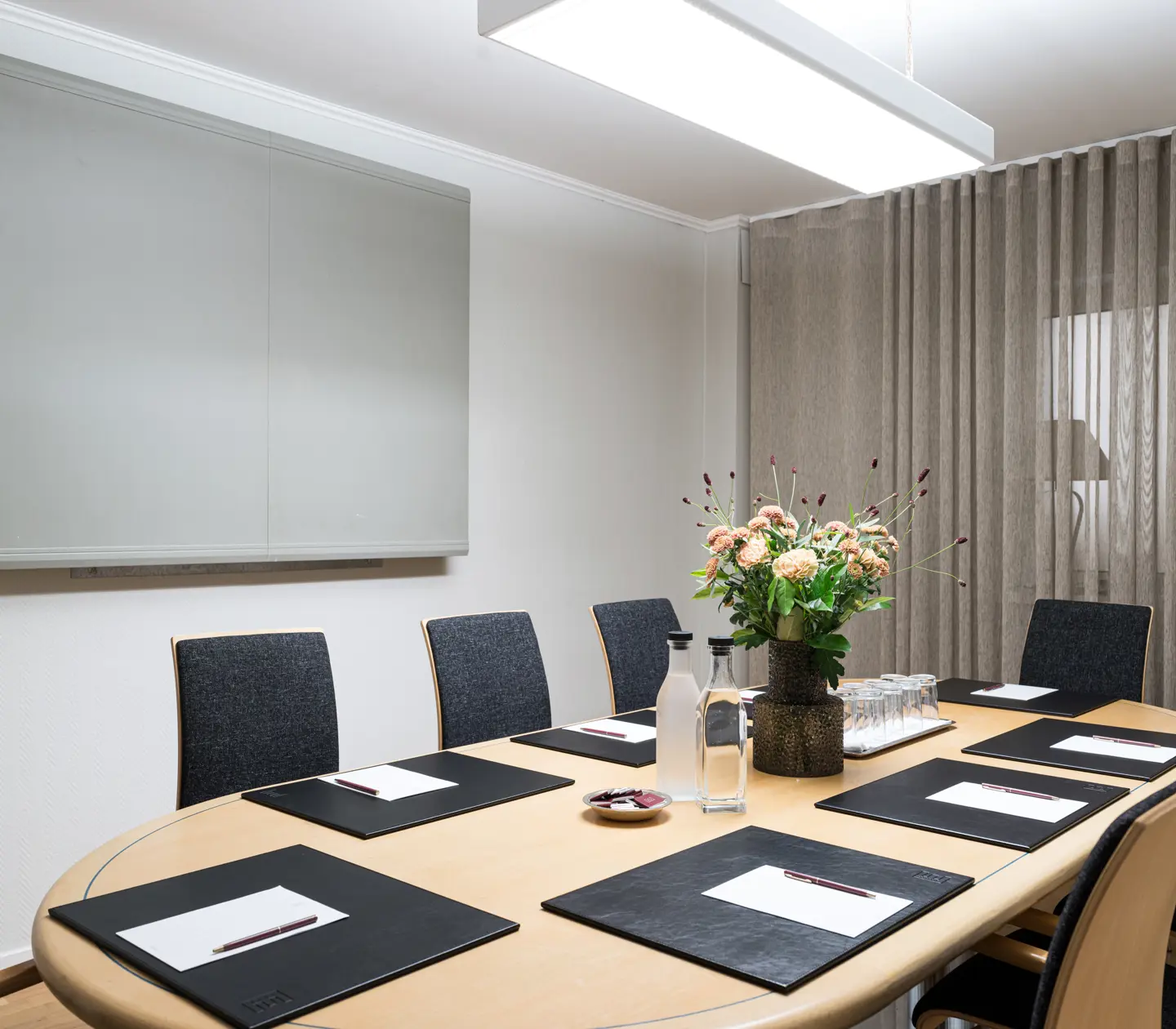 Konferensrum med oval träbord, svarta stolar och blommor i vas. Bordet är dukat med papper, pennor och vattenflaskor.