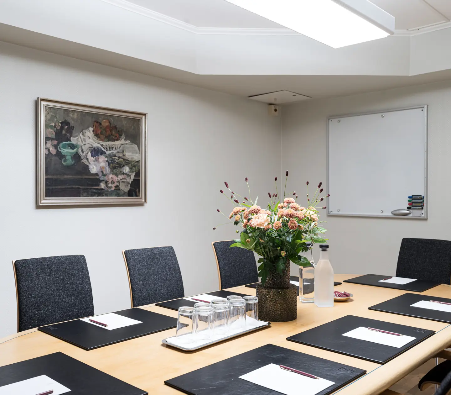 Konferensrum med stilfull inredning, ett avlångt bord omgiven av svarta stolar, blomsterarrangemang som mittpunkt, och anteckningsblock upplagda vid varje plats. En tavla och skrivtavla pryder väggarna, vilket skapar en inspirerande arbetsmiljö.