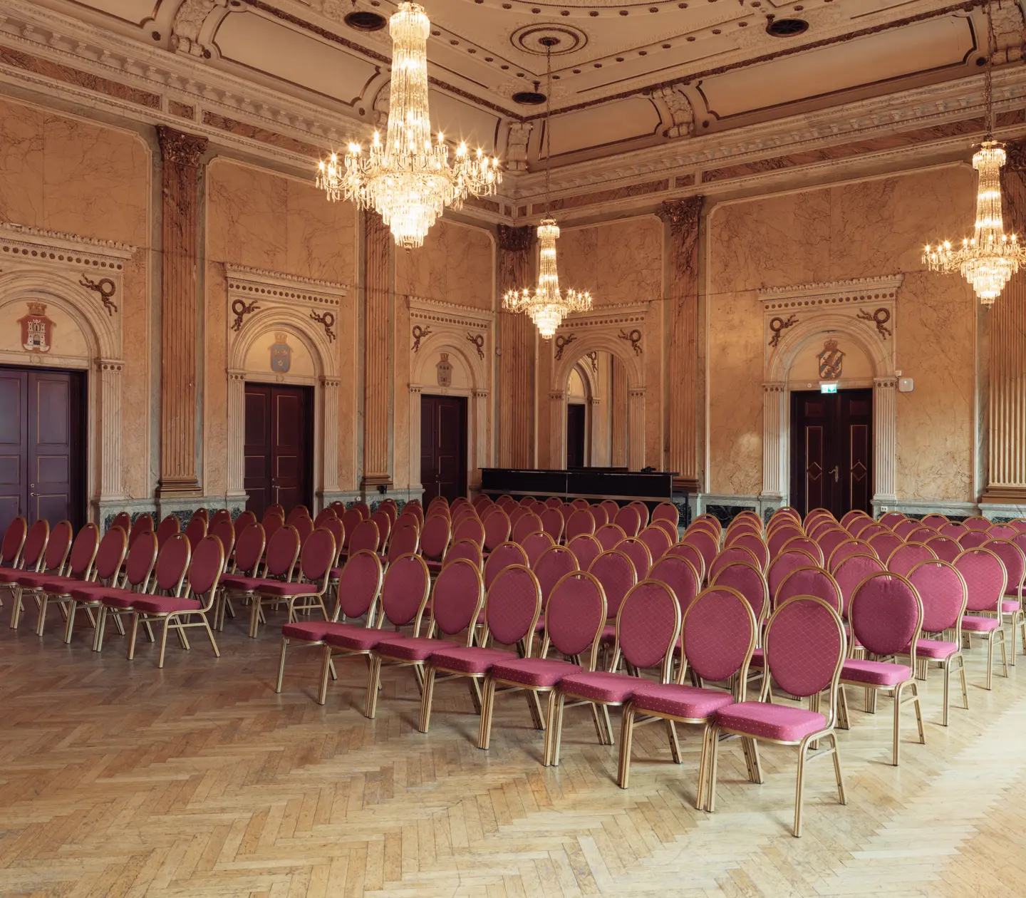 Bild på en elegant bankettsal med historisk arkitektur. Salen har vackra väggpaneler och dekorerade dörrar, med stora kristallkronor hängande från taket. Rader av stolar med röd klädsel och gyllene ramar är placerade på ett trägolv i fiskbensmönster.