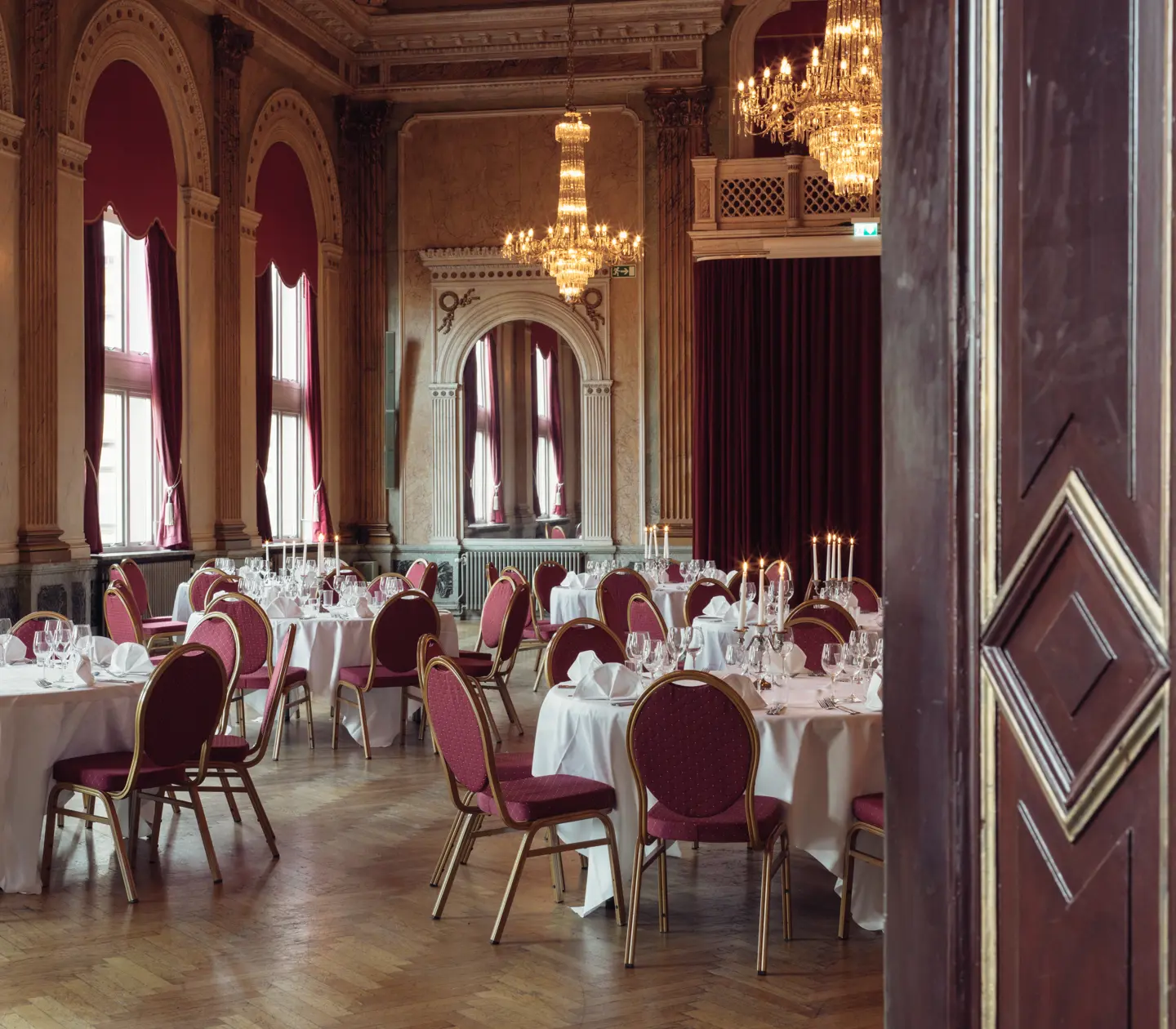 Elegant balsal med klassisk inredning, kristallkronor och runda, dukade bord med röda stolar.