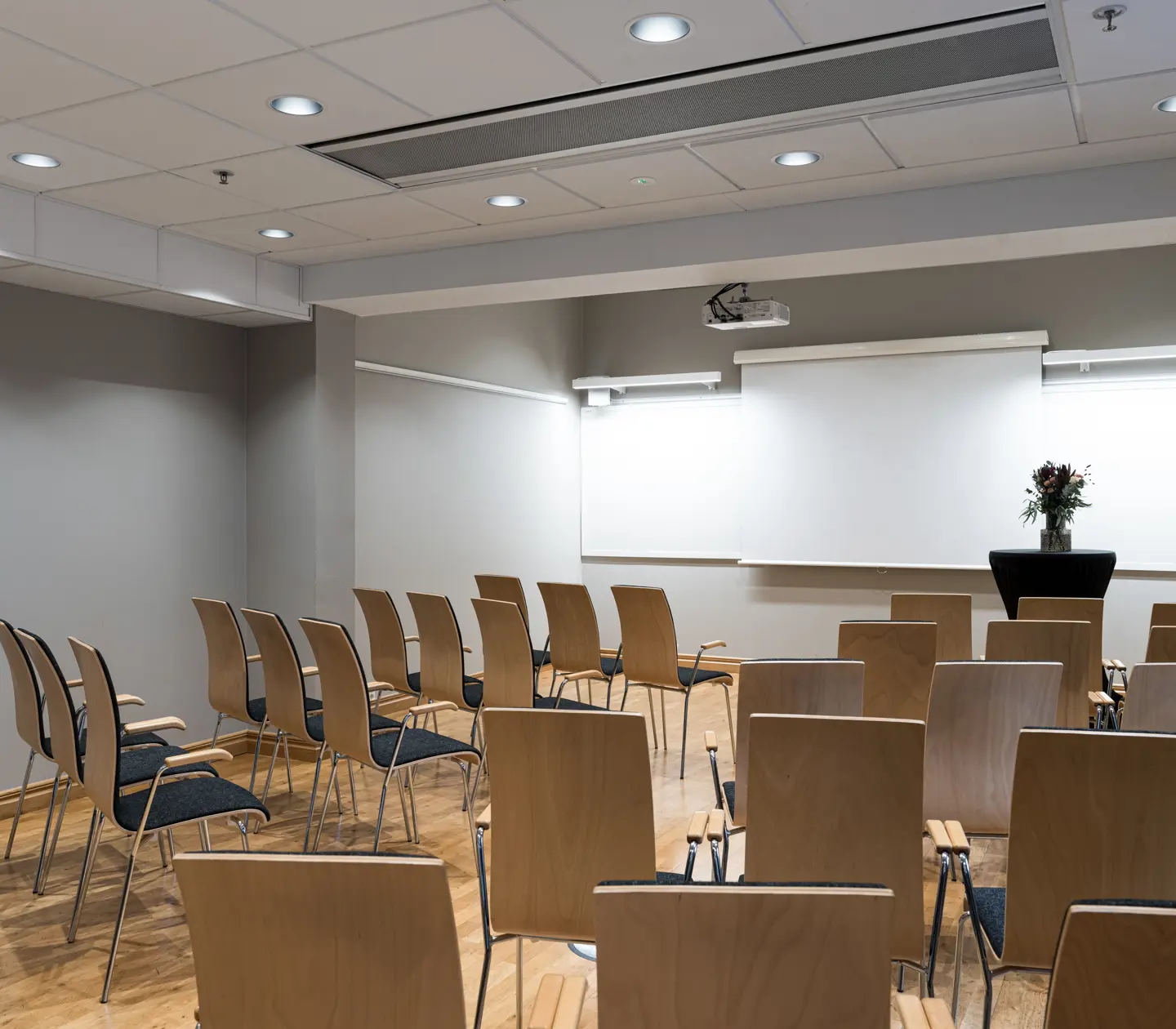 Konferensrum med uppställda stolar i rader, projektorduk och blombukett i hörnet.