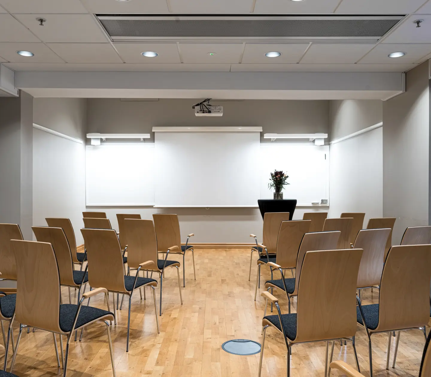Konferensrum med rad av trästolar, projektor och blädderblock på väggen, ljus interiör med blombukett på podium. Perfekt för möten och presentationer.