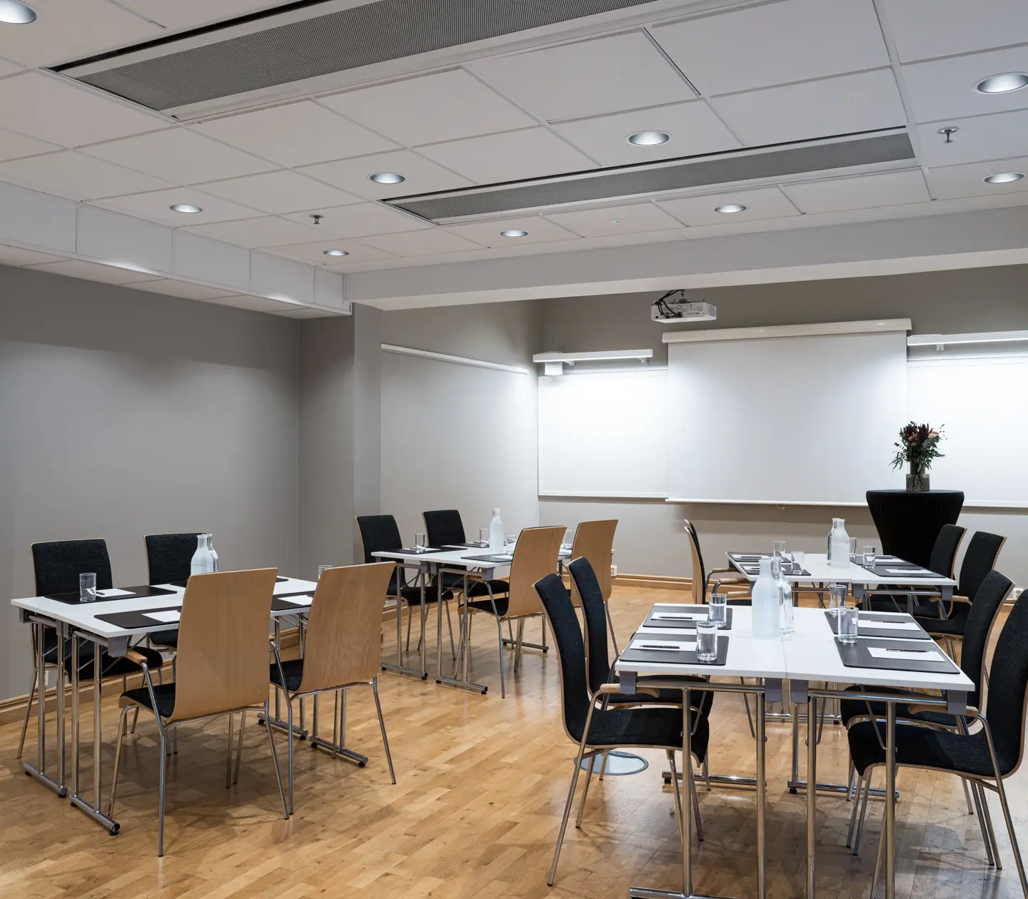 Konferensrum med arrangemang av stolar och bord, förberett för möte eller presentation, med projektorduk och blommor på ett podium.