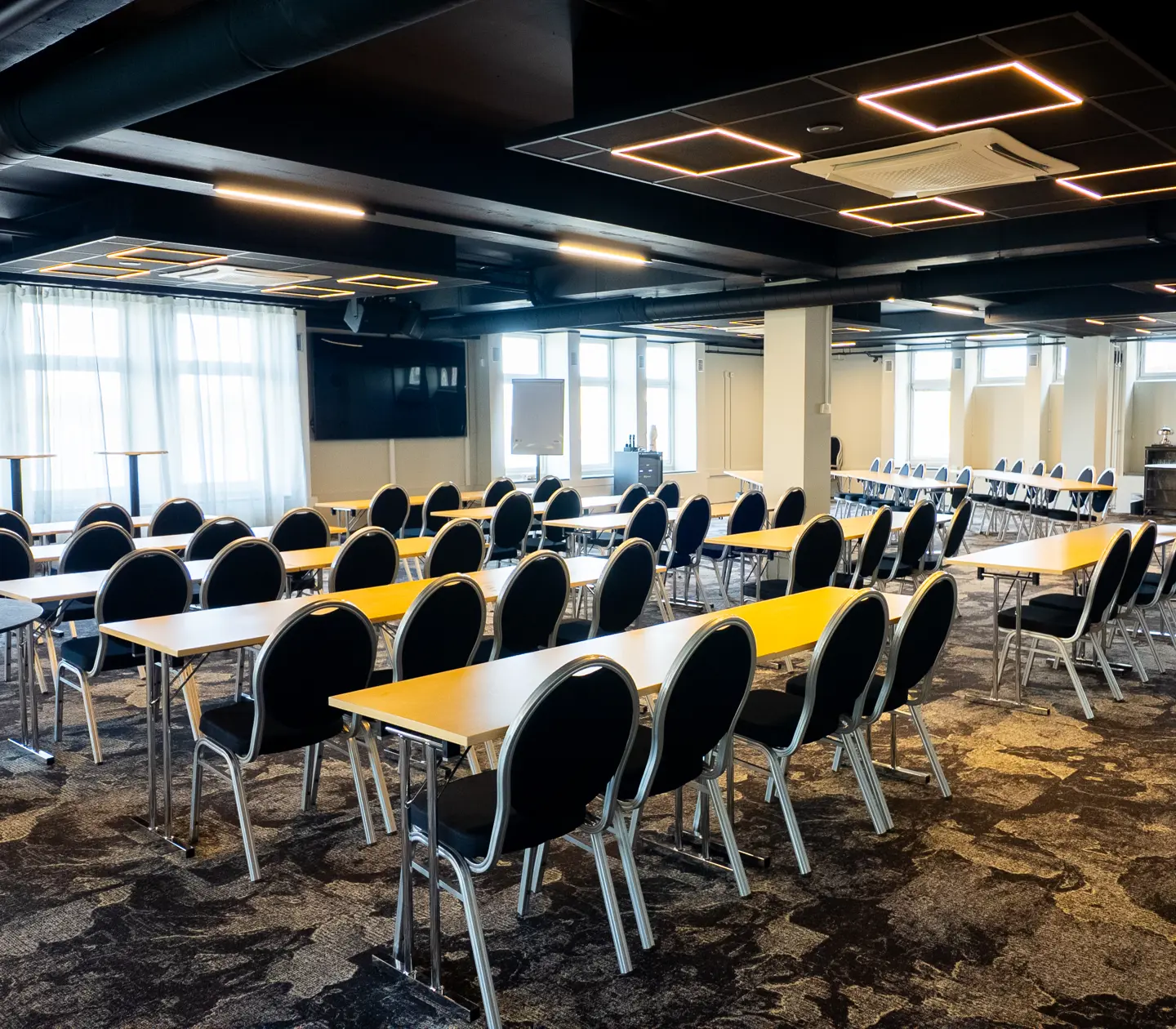 Spacious conference room with rows of tables and chairs, modern lighting, and large windows allowing natural light.