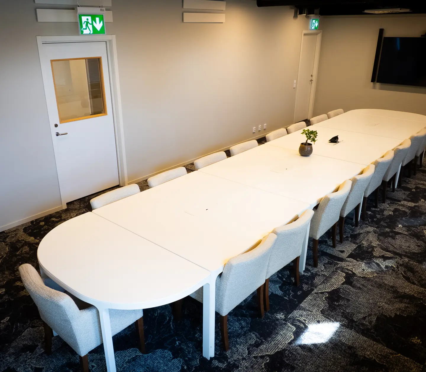 Modern conference room with a long white table surrounded by white chairs, featuring a wall-mounted screen and contemporary carpet flooring.