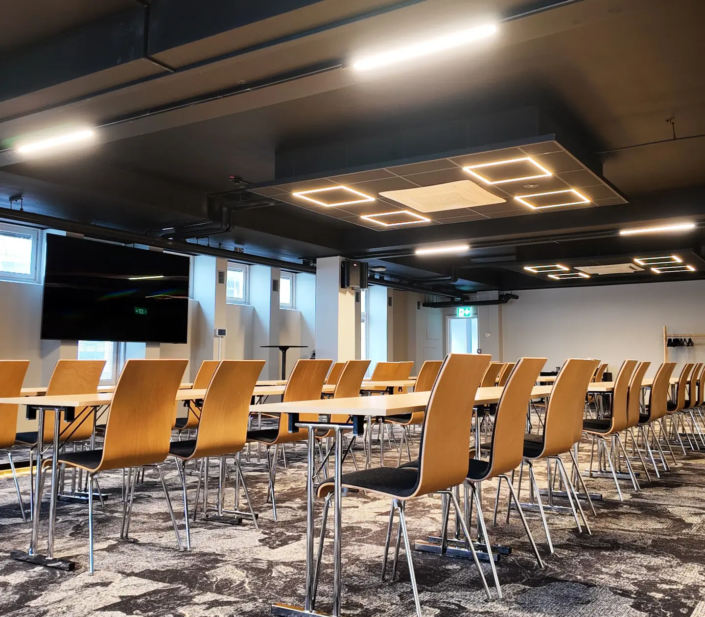 Modern conference room with rows of wooden chairs and tables, featuring a large screen and contemporary ceiling lights.