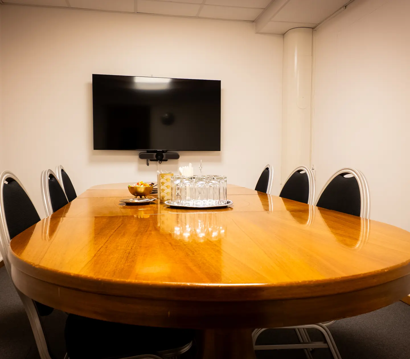 Modern conference room with a wooden oval table, six black chairs, a wall-mounted flat-screen TV, and a set of glasses and snacks in the center.