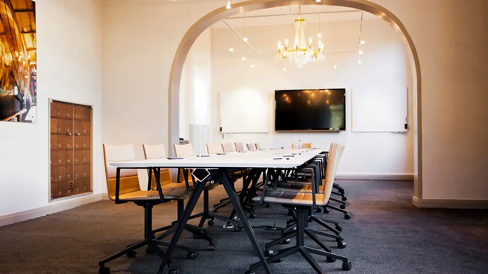 Modern conference room with long table, wooden chairs, large screen, chandelier, and arched entryway.