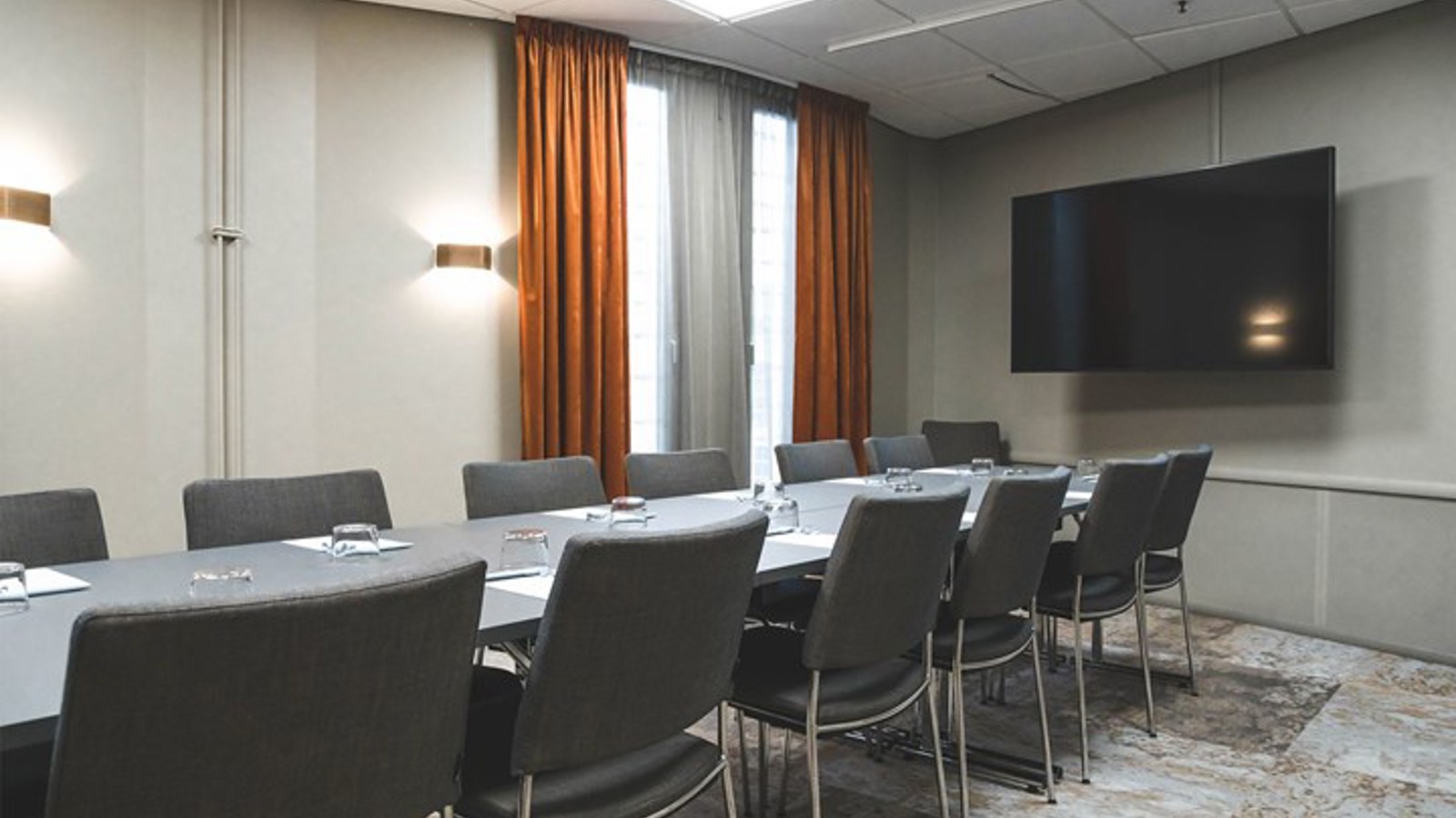 Konferensrum med styrelsesittning, grå stolar, grått bord, tv, rostbruna gardiner