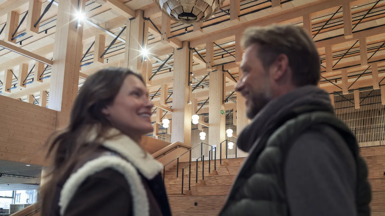 A man and woman smiling at each other inside a modern wooden architecture building with exposed beams and a large light fixture.